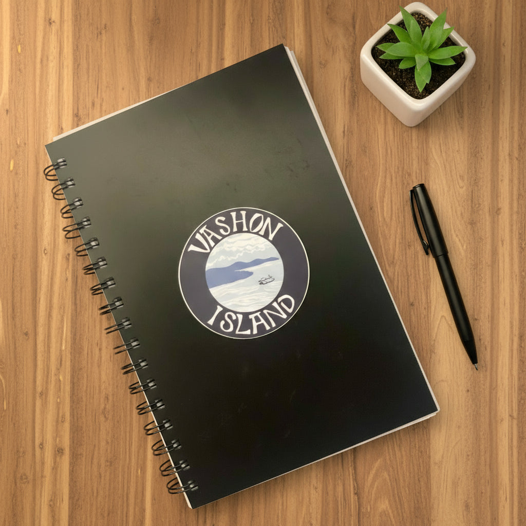 Black notebook with 'Vashon Island' logo on a wooden surface with a pen and plant.
